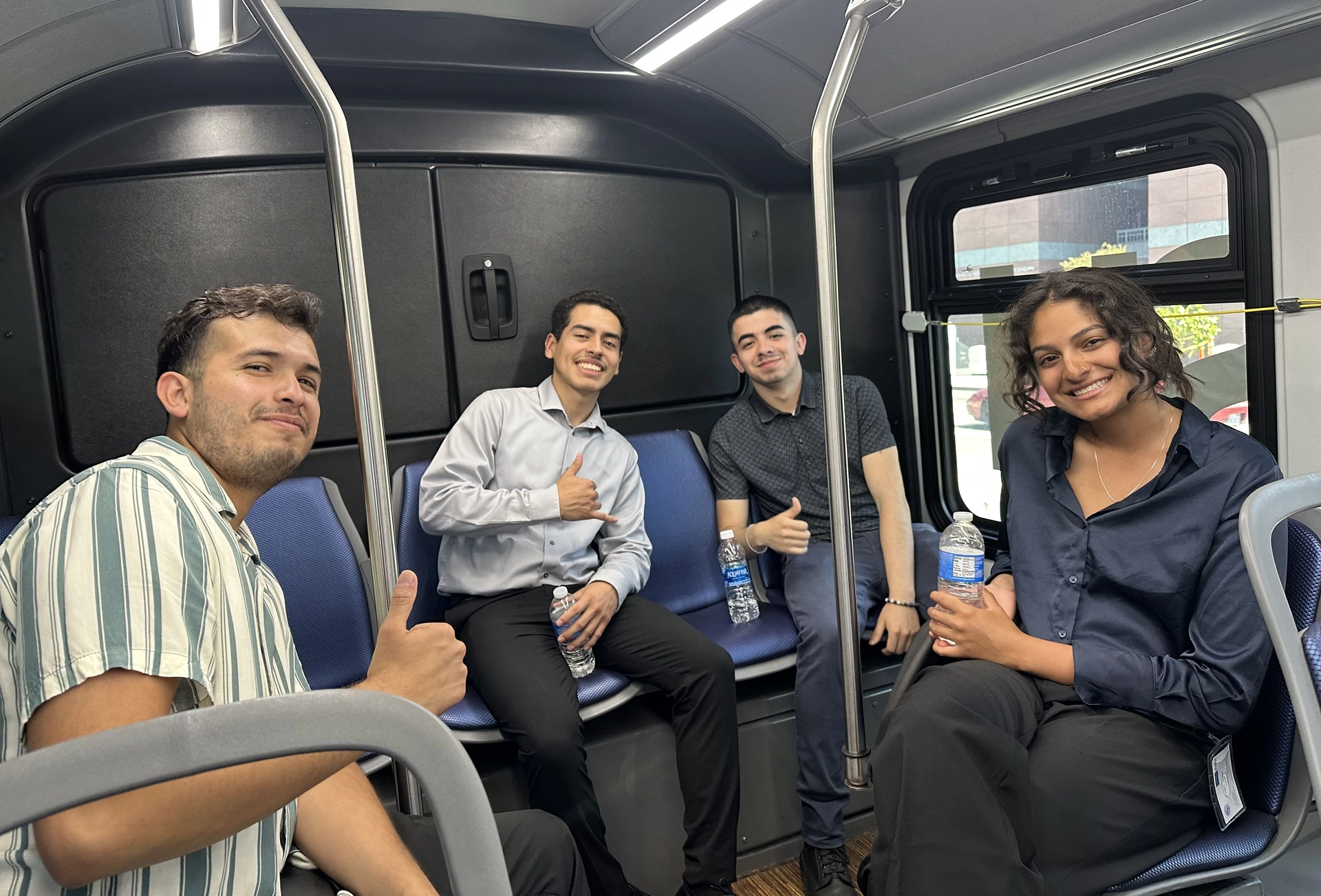 Four young interns holding water bottles, smiling, and giving a thumbs up while riding public transportation.