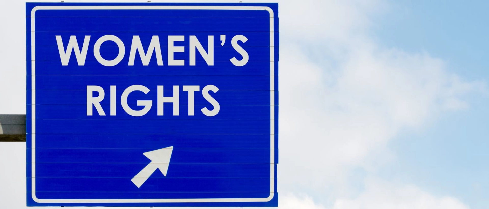 Stock photo of a dark blue freeway sign with the words, "Women's Right's" and an arrow in white. The sign is front of a blue sky with clouds.