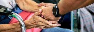 Close up shot of an older person in a metal chair holding the hands of a younger person.